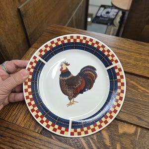 May include: A white ceramic plate with a rooster illustration. The plate has a red, white, and blue checkered border. The rooster is brown, red, and blue. The plate is on a wooden surface.