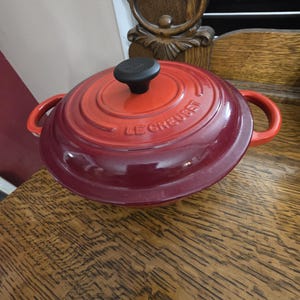 May include: A red and burgundy Le Creuset Dutch oven with a black knob and side handles. The lid has the brand name embossed on it. The cookware is on a wooden surface.