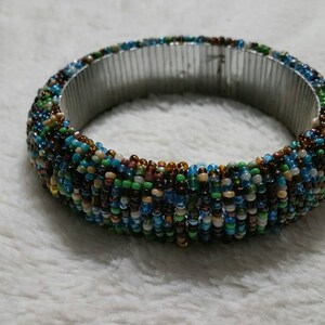 Op de afbeelding: Een kralen armband met een zilveren metalen band. De kralen zijn veelkleurig, waaronder bruin, blauw, groen en wit.