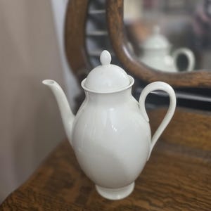Puede incluir: Una tetera de porcelana blanca con tapa, boquilla y asa. La tetera tiene un cuerpo redondeado y se asienta sobre una pequeña base. El fondo incluye una superficie de madera y un reflejo borroso en un espejo.