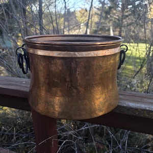 Puede incluir: Una maceta de metal color cobre con un cuerpo ancho y redondeado y un borde ligeramente ensanchado. Tiene dos asas negras y curvas a cada lado. La maceta está sobre una barandilla de madera, con un fondo borroso de árboles y cielo.