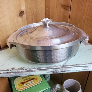 Op de afbeelding: Een vintage zilvermetalen serveerschaal met deksel. De schaal heeft een gehamerde textuur op de kom en een decoratieve handgreep op het deksel. De schaal staat op een lichtgroene plank.