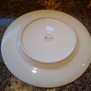 May include: White ceramic dinner plate with a raised rim. The bottom of the plate has a small, round sticker with the text "Franciscan Fine China" printed in black.