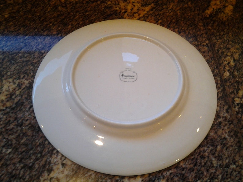 May include: White ceramic dinner plate with a raised rim. The bottom of the plate has a small, round sticker with the text "Franciscan Fine China" printed in black.