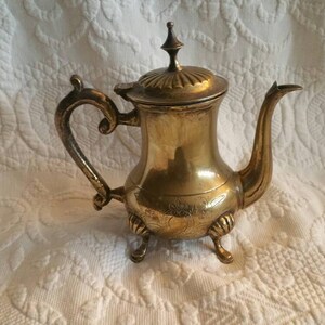 May include: A gold-colored metal teapot with an ornate design and a lid. The teapot has a curved spout and a handle with a decorative design. The teapot is sitting on a white surface.