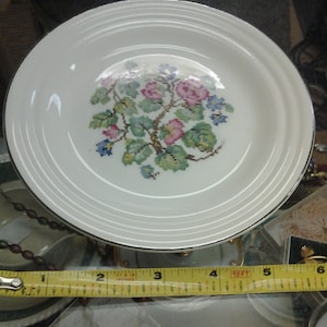May include: A white ceramic plate with a floral design featuring pink, blue, and green flowers. The plate has a gold rim and is approximately 7 inches in diameter.