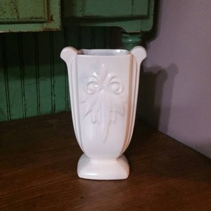 May include: A white ceramic vase with a decorative leaf design. The vase has a square base and a wide mouth.