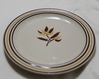May include: A white ceramic dinner plate with a brown and yellow floral design. The plate has a brown rim with three thin yellow stripes.