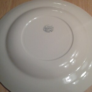 Puede incluir: Plato de cer&aacute;mica blanco con un borde elevado y un dise&ntilde;o circular en la parte inferior. El plato tiene una marca azul tenue que dice "St. Francis, Made in England".