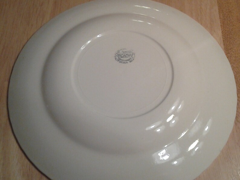 Puede incluir: Plato de cer&aacute;mica blanco con un borde elevado y un dise&ntilde;o circular en la parte inferior. El plato tiene una marca azul tenue que dice "St. Francis, Made in England".