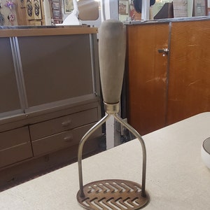 Puede incluir: Un machacador de patatas de plata con un mango de madera. El machacador tiene una base redonda perforada y un mango en forma de Y.