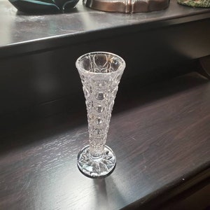 Può includere: Un vaso di vetro trasparente per un solo bocciolo, con un motivo testurizzato a forma di diamante. Il vaso ha un corpo alto e sottile e una base svasata.
