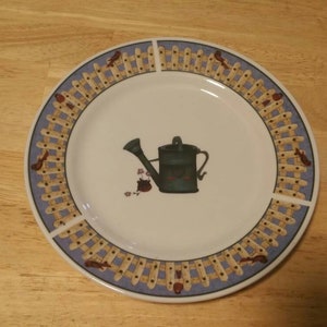 May include: A white ceramic plate with a blue and yellow border featuring a stylized image of a watering can with flowers. The border design is a repeating pattern of a picket fence.