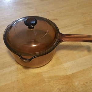 Puede incluir: Un cazo de cristal de color ámbar translúcido con una tapa circular de cristal a juego. La olla cuenta con un mango largo de cristal integrado que se extiende por un lado y un pequeño pico vertedor en el borde opuesto al mango.
