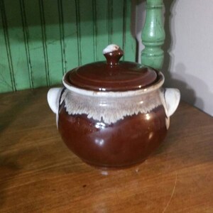 May include: A brown ceramic crock pot with a white lid and white accents. The crock pot has a glazed finish and a rounded shape.