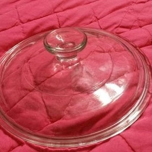 Puede incluir: Una tapa de vidrio transparente con un pomo en la parte superior. La tapa está sobre una superficie de tela roja.