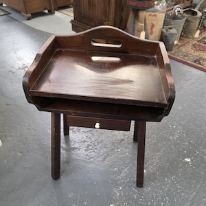 May include: A dark brown wooden side table with a tray top and a small drawer. The table has a handle cut out of the top and is supported by four legs. The drawer has a white knob. The table is likely vintage or antique.