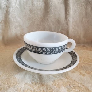 May include: A white teacup and saucer with a black and grey leaf design. The cup and saucer are made of ceramic.