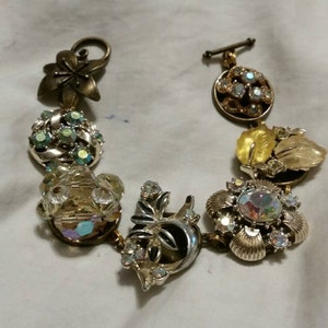 May include: A gold-tone bracelet featuring a selection of vintage-style charms, including a flower, a leaf, and several jeweled buttons.