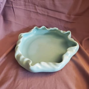 Op de afbeelding: Een lichtblauwe keramische kom met een golvende, gekrulde rand. De kom is leeg en heeft een gladde, glanzende afwerking.