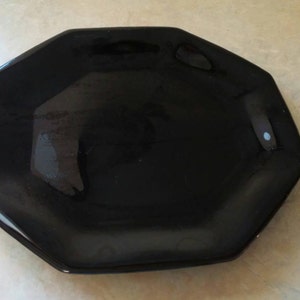 May include: A glossy black octagonal plate with a smooth, reflective surface and defined edges. The plate features a solid dark color throughout its entire structure, presenting a simple geometric shape with eight distinct sides and a flat center.