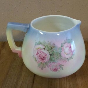 May include: A white ceramic jug with a blue, yellow and pink handle. The jug has a gradient design of blue, pink and white. It features a floral design with pink roses and green leaves.