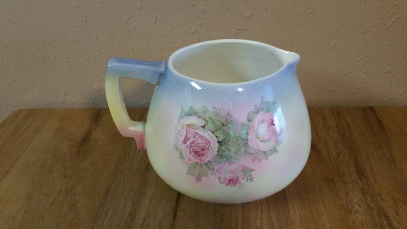 May include: A white ceramic jug with a blue, yellow and pink handle. The jug has a gradient design of blue, pink and white. It features a floral design with pink roses and green leaves.