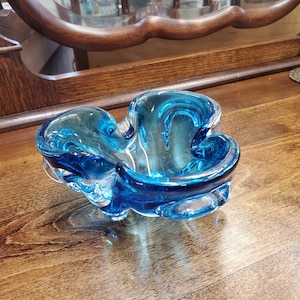 Può includere: Un posacenere in vetro blu con un design ondulato e organico. Il posacenere è realizzato in vetro trasparente con una tinta blu e ha una forma asimmetrica unica.