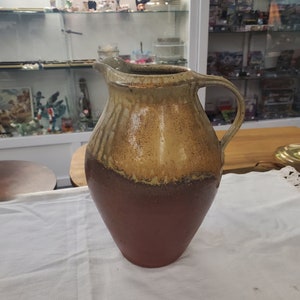 Può includere: Un'anfora in ceramica marrone e beige con un'ampia apertura e un manico. L'anfora ha un aspetto rustico e fatto a mano.