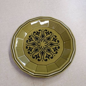 Könnte beinhalten: Zwei olivgrüne Keramikplatten übereinander gestapelt. Die Teller haben eine achteckige Form mit einem schwarzen floralen Design in der Mitte. Die Teller sind einfarbig mit glänzendem Finish. Geschätzter Durchmesser: 20-25 cm.