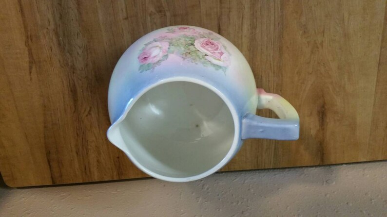 May include: A blue and white ceramic jug with a floral design featuring pink roses. The jug has a handle and a spout.