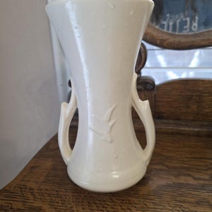 Könnte beinhalten: Cremefarbene Keramikvase mit ausgestelltem Oberteil und zwei geschwungenen Griffen. Die Vase hat ein dezentes geprägtes Design und steht auf einer Holzoberfläche. Die Vase hat eine glänzende Oberfläche und eine klassische, elegante Form.