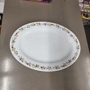 May include: An oval, white ceramic serving platter with a floral border. The border features a repeating pattern of small blue, red, and yellow flowers with green leaves. The platter is empty and sits on a neutral-toned surface.