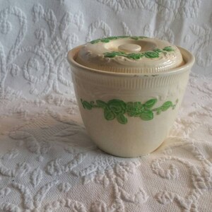 May include: A white ceramic jar with a lid, decorated with green floral designs. The jar has a smooth, rounded shape and a slightly flared rim.