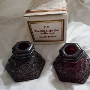 May include: Two dark red glass candle holders with a decorative design. The holders are part of the Avon 1876 Cape Cod Collection.