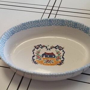 May include: A white oval baking dish with a blue and white dotted pattern around the rim. The center of the dish features a colorful illustration of a small house with flowers and ribbons.