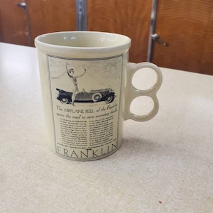 Puede incluir: Una taza de cerámica blanca con doble asa. La taza presenta una ilustración en blanco y negro de un automóvil antiguo con el texto "The AIRPLANE FEEL of the Franklin opens the road to new motoring thrills" y "FRANKLIN 1929".
