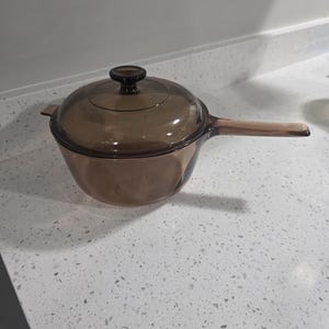 Puede incluir: Un cazo de cristal marrón translúcido con su tapa a juego. La olla cuenta con un mango largo integrado en un lado y un pequeño pomo circular en el centro de la tapa. El cristal tiene un acabado liso y pulido con un tono ámbar profundo.