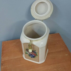 K&ouml;nnte beinhalten: Wei&szlig;er Keramikbeh&auml;lter mit Deckel, mit einem Blumendesign aus Blaubeeren und anderen Beeren. Der Beh&auml;lter hat einen goldfarbenen Verschluss und ist offen, wodurch das Innere sichtbar wird. Er steht auf einer Holzoberfl&auml;che.