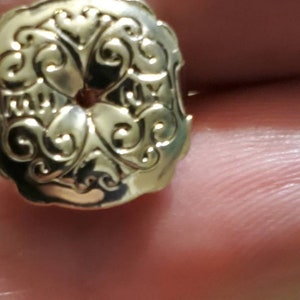 May include: A gold-toned metal button with a decorative, ornate design. The button has a circular hole in the centre.
