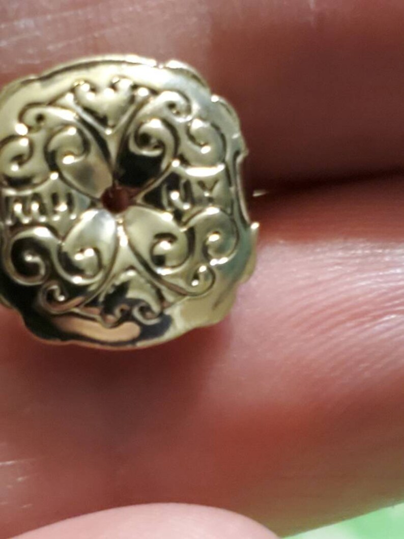 May include: A gold-toned metal button with a decorative, ornate design. The button has a circular hole in the centre.