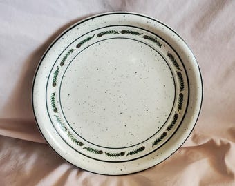 Op de afbeelding: Witte keramische dinerbord met een groene en zwarte rand met een herhalend patroon van dennennaalden en dennenappels.