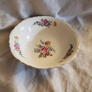 Puede incluir: Un tazón de cerámica blanca con un borde dorado y un patrón floral. El tazón presenta flores rosas, amarillas y azules con hojas verdes.