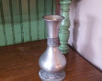 May include: A silver pewter vase with an ornate floral design sits on a wooden table. The vase has a round base and a narrow neck.