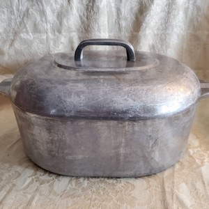 Op de afbeelding: Een ovale aluminium kookpot met een deksel. De pot heeft een handvat op het deksel en twee handvatten aan de zijkanten. Het metaal heeft een versleten, geborsteld uiterlijk, wat duidt op leeftijd en gebruik. Een klassiek keukengerei.