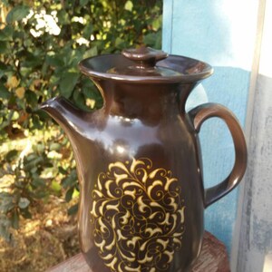 May include: A brown ceramic coffee pot with a lid and a gold floral design. The pot is sitting on a wooden surface.