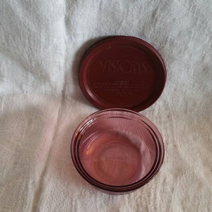 Può includere: Un contenitore di vetro rosso con un coperchio abbinato. Il coperchio ha la scritta "VISIONS" stampata in lettere bianche. Il contenitore è vuoto.