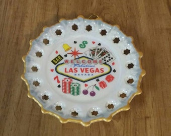 May include: White ceramic plate with a gold rim and a Las Vegas design. The plate features a welcome to fabulous Las Vegas sign, playing cards, dice, cherries, and a bell. The plate has a scalloped edge with cutouts.
