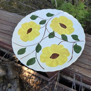 May include: A white ceramic plate with a scalloped edge, hand-painted with three large yellow flowers, brown centers, and green leaves. The flowers are connected by brown stems. The plate is resting on a wooden surface.