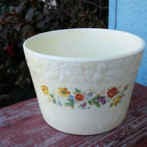 May include: A white ceramic planter with a floral design. The planter has a raised rim and a scalloped edge. The flowers are in shades of yellow, orange, and blue.